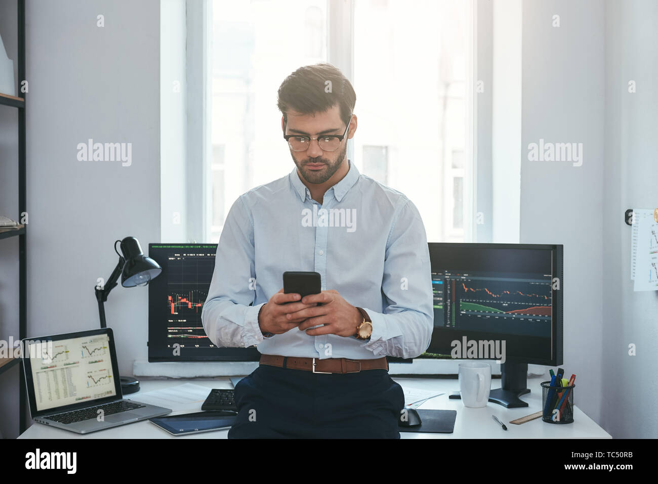 Moderne Technologien. Erfolgreiche junge Unternehmer in formalen Verschleiß und Brillen mit Smartphone, während im Büro vor den Bildschirmen mit den Charts. Forex Markt. Geschäftskonzept. Investitionen Konzept Stockfoto