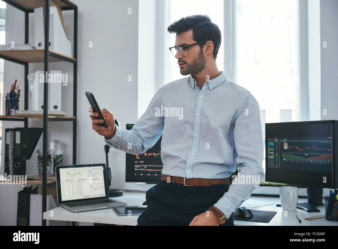 Börse. Konzentrierte junge Unternehmer oder Unternehmer, die in förmlichen Verschleiß und Brillen ist der Handel berichtet über sein Smartphone für Investitionen Zweck während in seinem Büro arbeiten. . Crypto Währungen. Geschäftskonzept. Investitionen Konzept Stockfoto