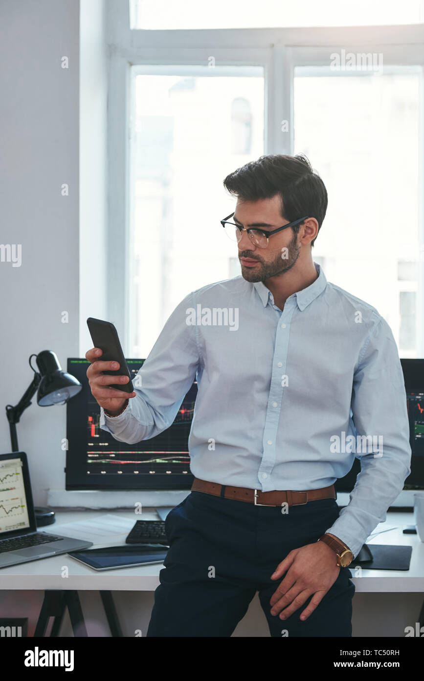 Erfolgreicher Geschäftsmann. Konzentrierte junge Unternehmer in formalen Verschleiß und Brillen ist der Handel Diagramme und Berichte auf seinem Smartphone für Investitionen Zweck während in seinem Büro arbeiten. Forex Markt. Handel Konzept. Investitionen Konzept Stockfoto