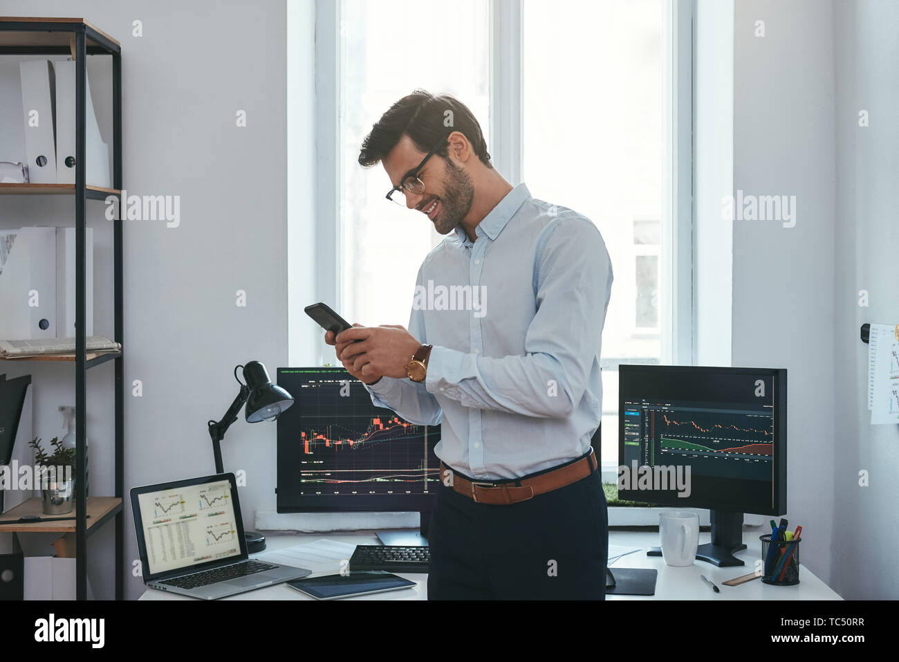 Modernes unternehmen. Glückliche und erfolgreiche Händler in formalen Verschleiß ist auf der Suche nach Handel Diagramme und Berichte auf seinem Smartphone für Investitionen Zweck und lächelnd, während in seinem Büro stehen. Forex Markt. Handel Konzept. Investitionen Konzept Stockfoto