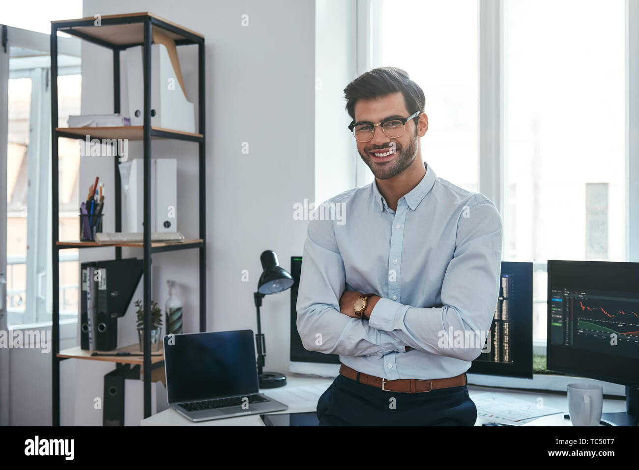 Gerne Händler. Fröhliche Geschäftsmann in formelle Kleidung und Brillen ist, halten die Arme verschränkt, lächelnd und mit Kamera, während im Büro stehen. Börsenmakler. Forex Markt. Handel Konzept Stockfoto