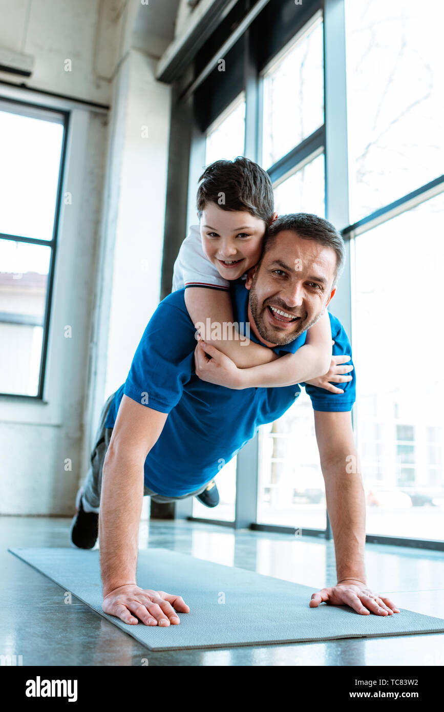 Gerne Vater tun Push up Übung mit Sohn auf dem Rücken im Fitnessstudio Stockfoto