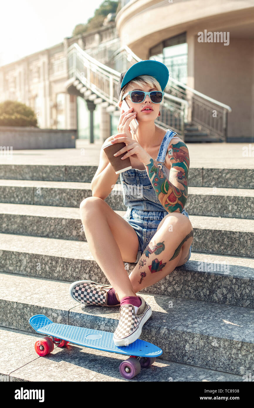 Junge alternative Mädchen skater tragen Cap und Sonnenbrille sitzen auf betontreppen Fuß auf Penny board Holding Tasse heißen Kaffee auf SMARTPHON Stockfoto