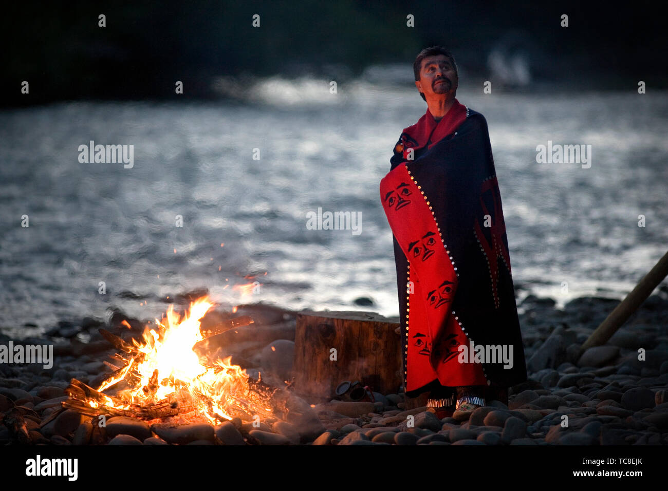Mitte - erwachsener Mann mit einer Decke um ihn, da er steht neben einem Lagerfeuer auf einem felsigen Strand Stockfoto
