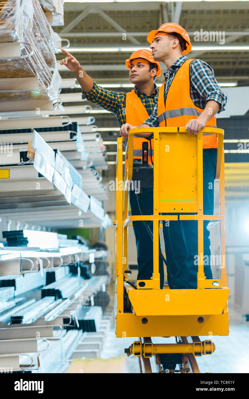 Ernsthafte indischer Arbeitnehmer zeigt mit dem Finger auf Baumaterialien beim Stehen auf scherenarbeitsbühne in der Nähe von Kollegen Stockfoto