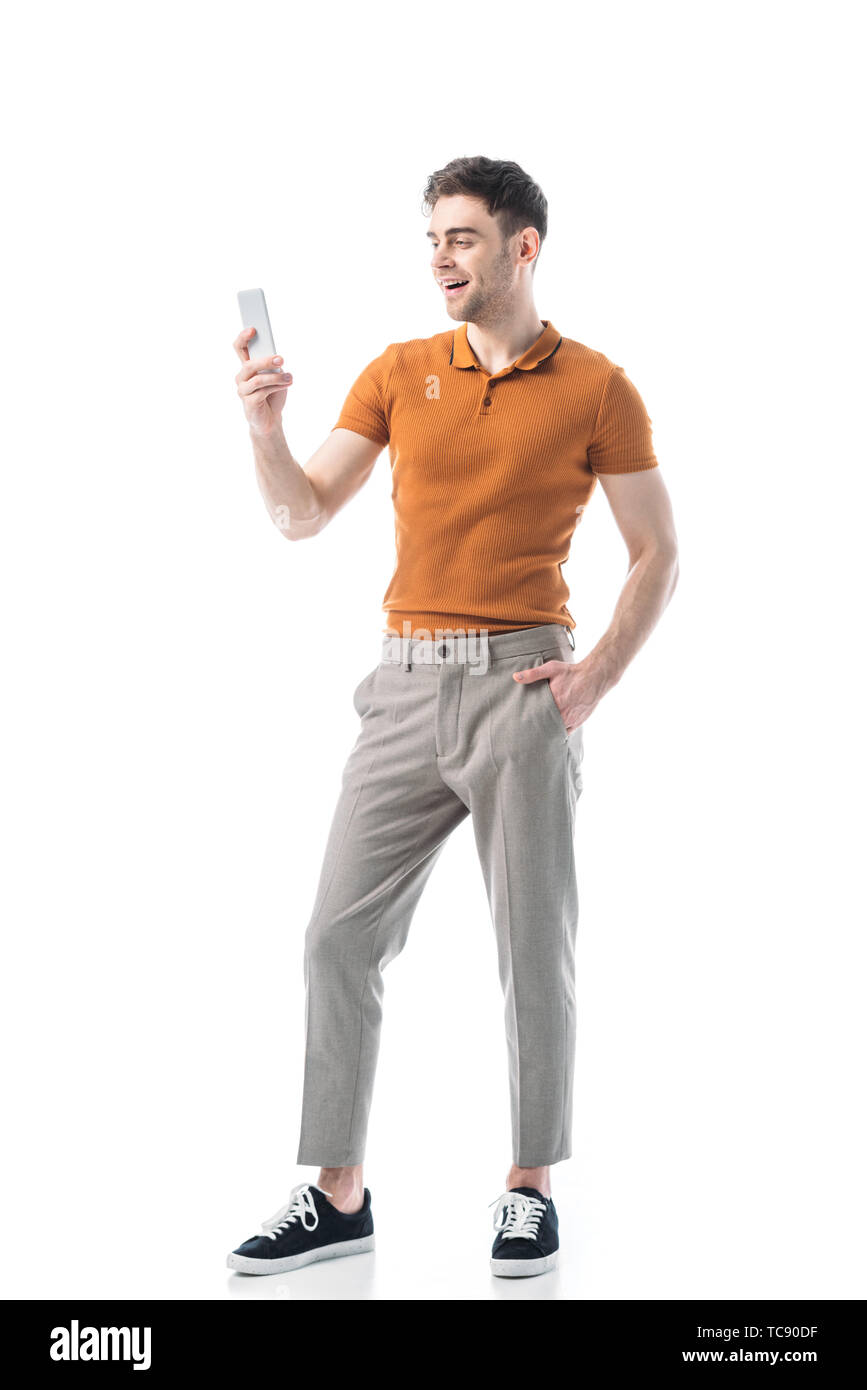 Gut aussehende fröhliche Mann mit Hand in der Tasche über Smartphone isoliert auf weißem Stockfoto