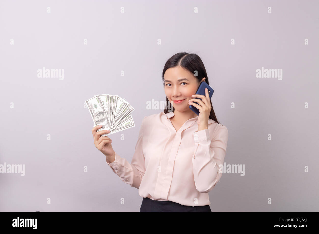 Asiatische schöne Frau holding Banknote Geld in der Hand und dem Handy in der anderen Hand auf grauem Hintergrund isoliert. kommerzielle Geschäft telefonisch anhand von quantitativen Simulatio Stockfoto
