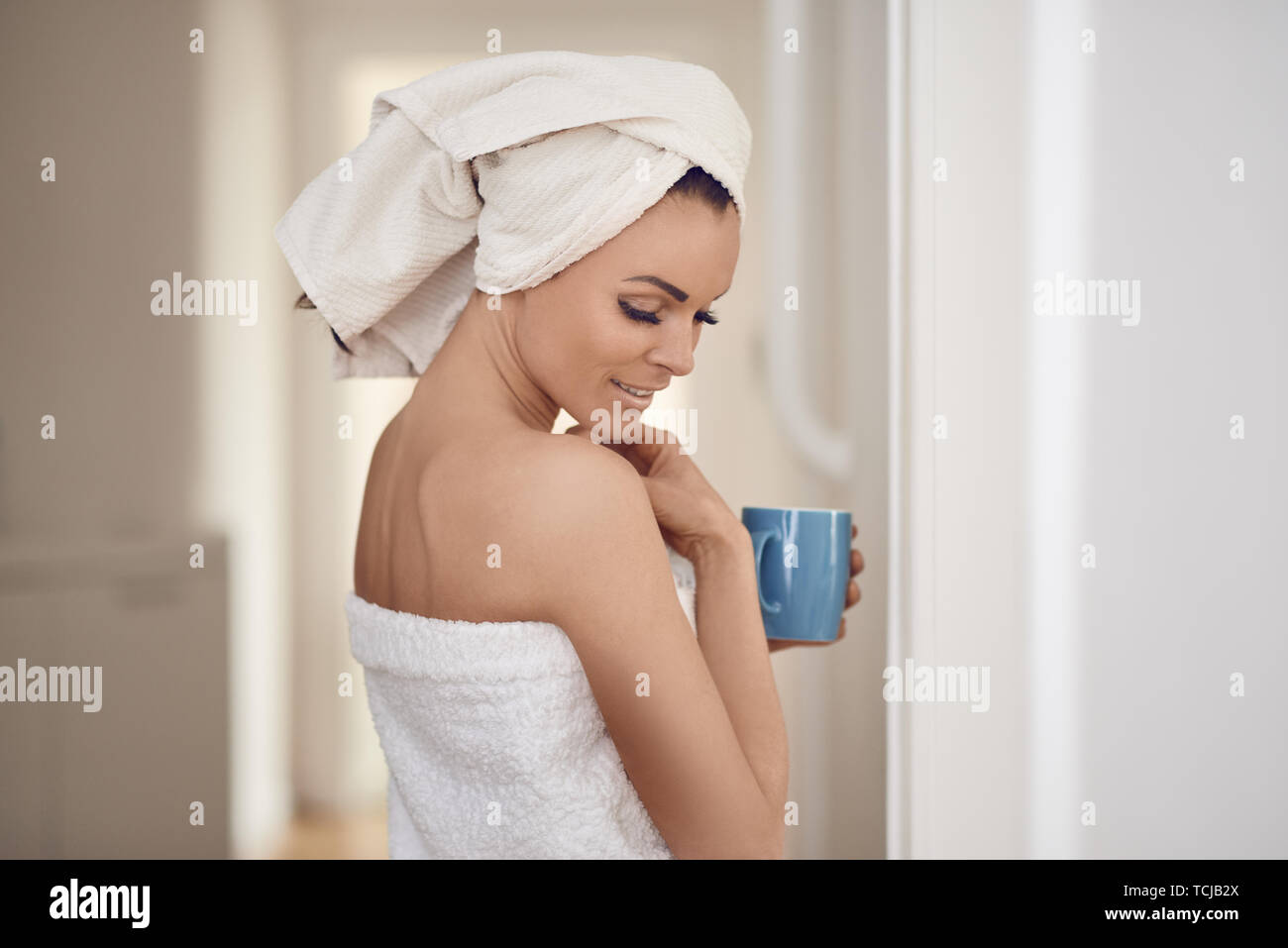 Hübsche Frau mittleren Alters in frische, saubere, weiße Handtücher um ihren Kopf und Körper gegen die Innenwand mit einer Tasse trinken in einem Con schiefen gewickelt Stockfoto