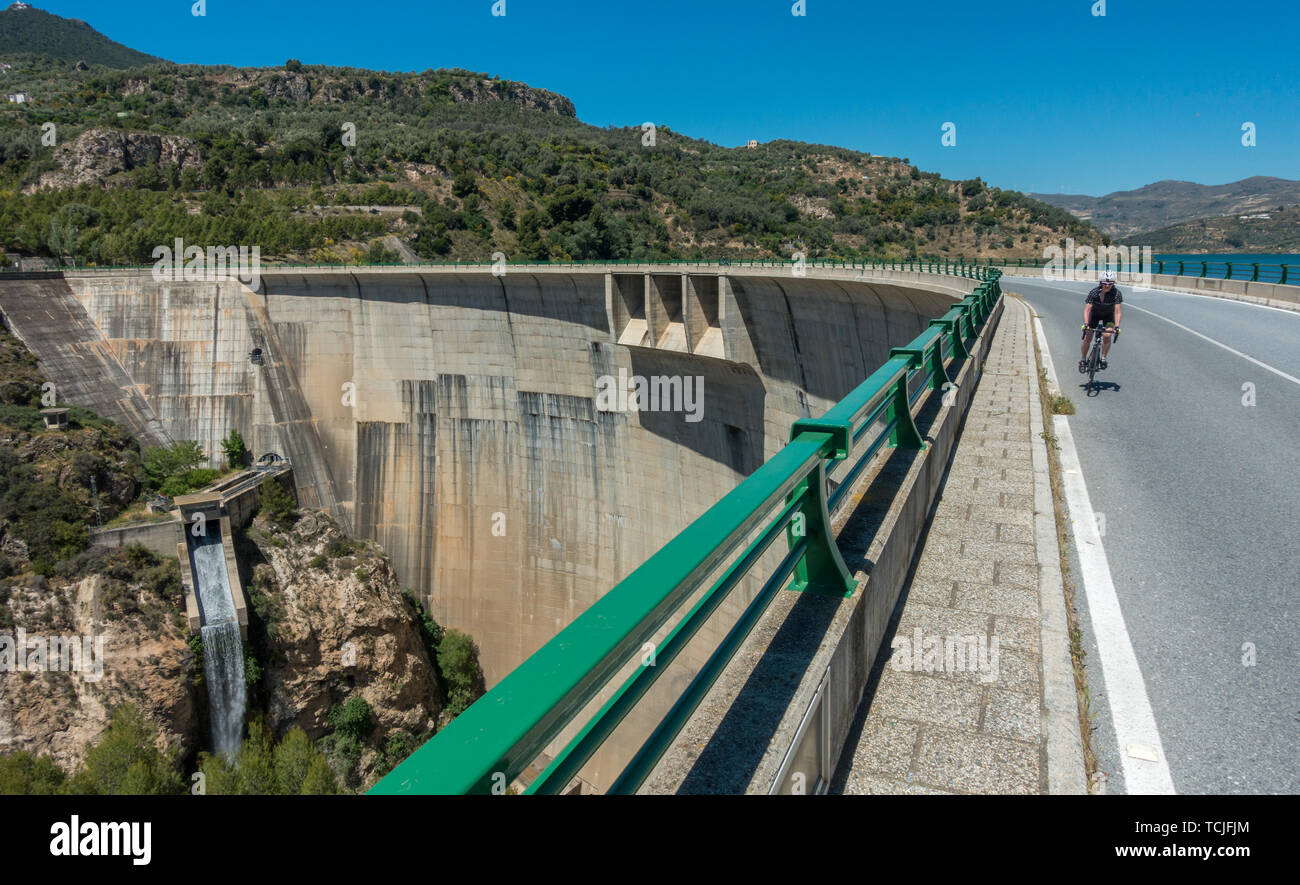 Weibliche Radfahrer reiten über den Damm am Stausee Embalse de Béznar mit dem Wasserfall, Presa de Béznar, im Hintergrund, in der Sierra Nevada, Spai Stockfoto
