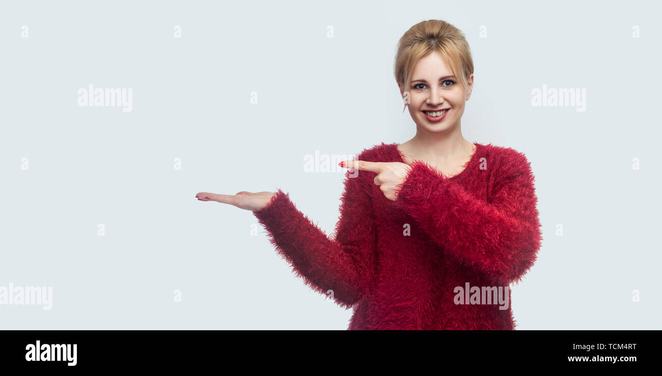 Portrait von glücklich Schöne junge blonde Frau in Rot Bluse stehen, zu Kamera mit toothy Lächeln suchen, zeigen auf etwas Holding an Hand. indo Stockfoto