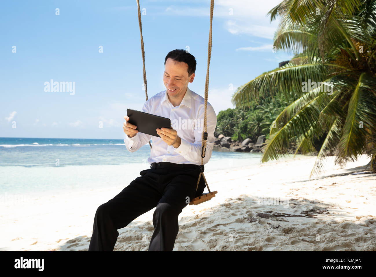 Mann sitzt auf Schaukel mit digitalen Tablet am Sandstrand in der Nähe der idyllischen Meer Stockfoto