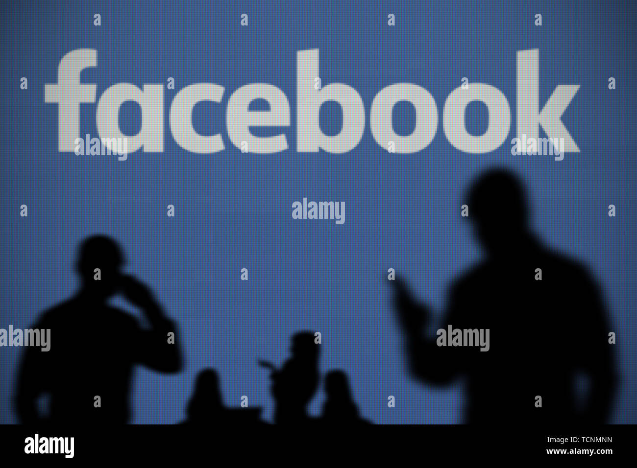 Das Facebook Logo auf einer LED-Bildschirm im Hintergrund, während eine Silhouette Person ein Smartphone verwendet im Vordergrund (nur redaktionelle Nutzung) Stockfoto