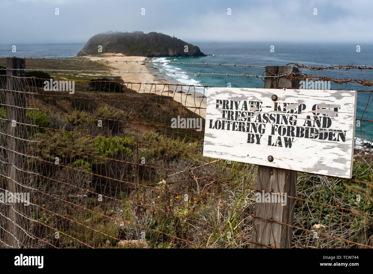 Eine verschlissene kein Übertreten Zeichen vor einem Zaun mit Stacheldraht, im Hintergrund ein wunderschöner Sandstrand und das Meer abstürzt. Stockfoto