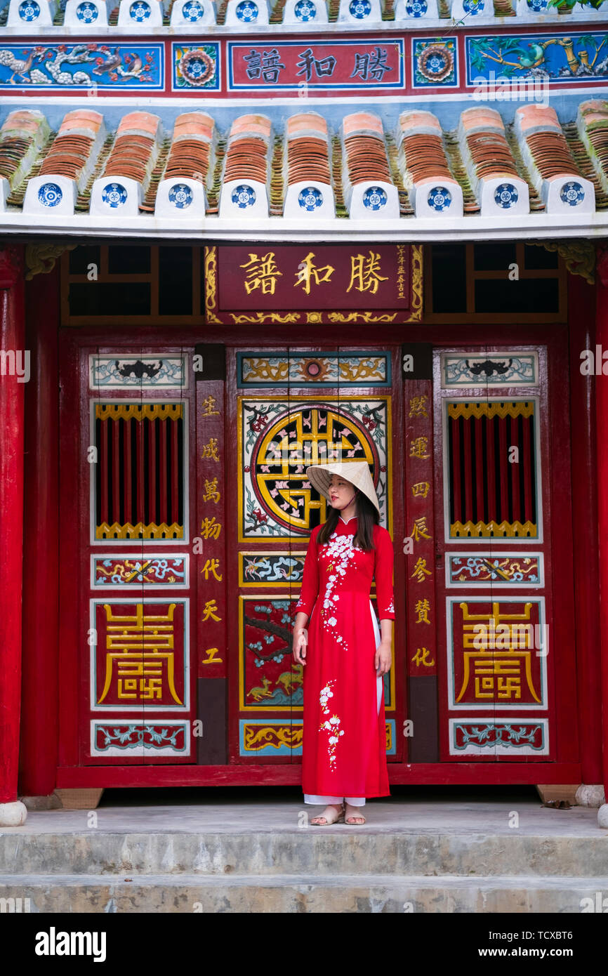 Eine junge Vietnamesin in einem traditionellen Ao Dai-Kleid und steht vor einem Tempel im historischen Stadtzentrum von Hoi an, Vietnam Stockfoto