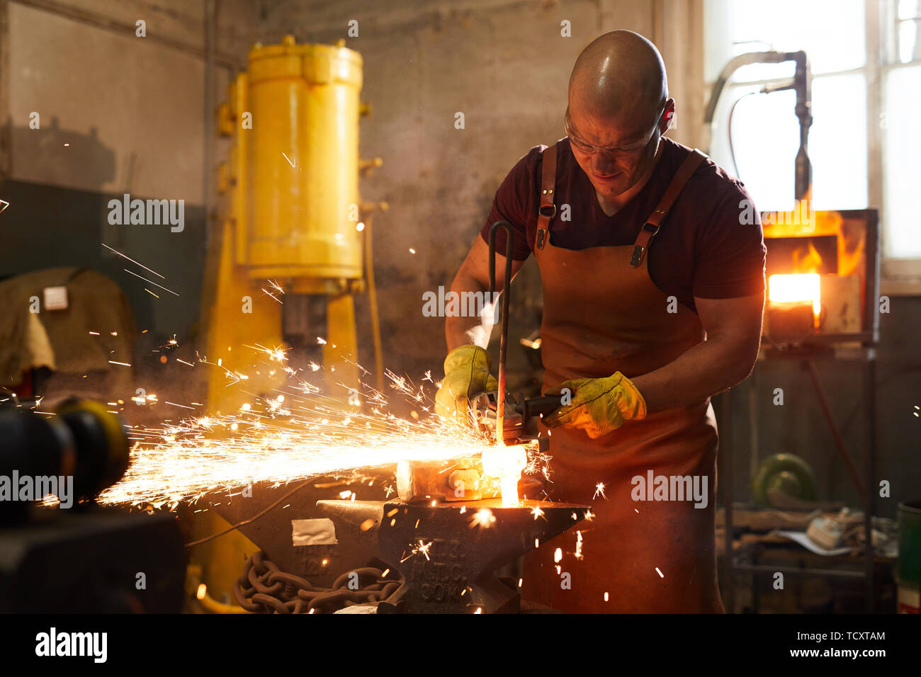 Stirnrunzelnd kahlen Schmied in Schürze schneiden beheizten Metall Stück mit Schleifer auf Amboss während der Arbeit in der Schmiede Stockfoto