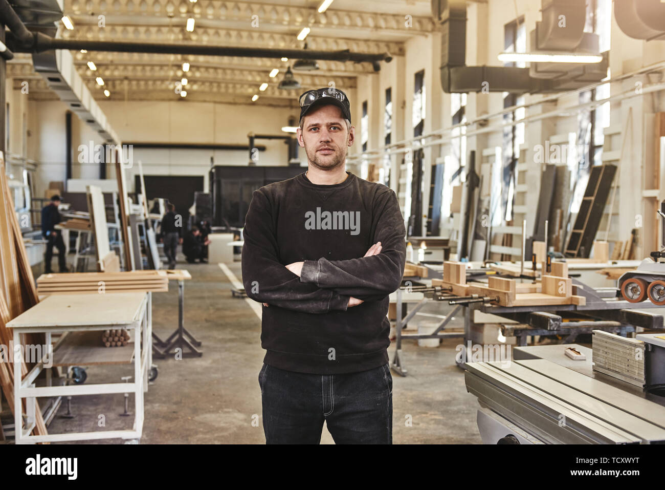 Kaukasische Mann stand mit verschränkten Armen und mit Blick auf die Kamera, während in der Möbelfabrik arbeiten Stockfoto