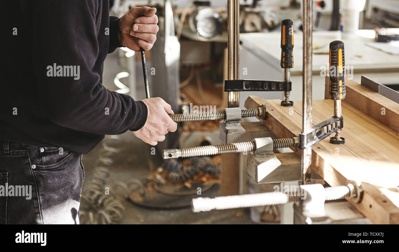 Handwerker mit Schraubzwingen fixieren zwei Stücke Holz und Eisen. Der Prozess der Herstellung Schreibtisch, Möbel. Arbeiter im Hintergrund. Horizontale Schuß Stockfoto