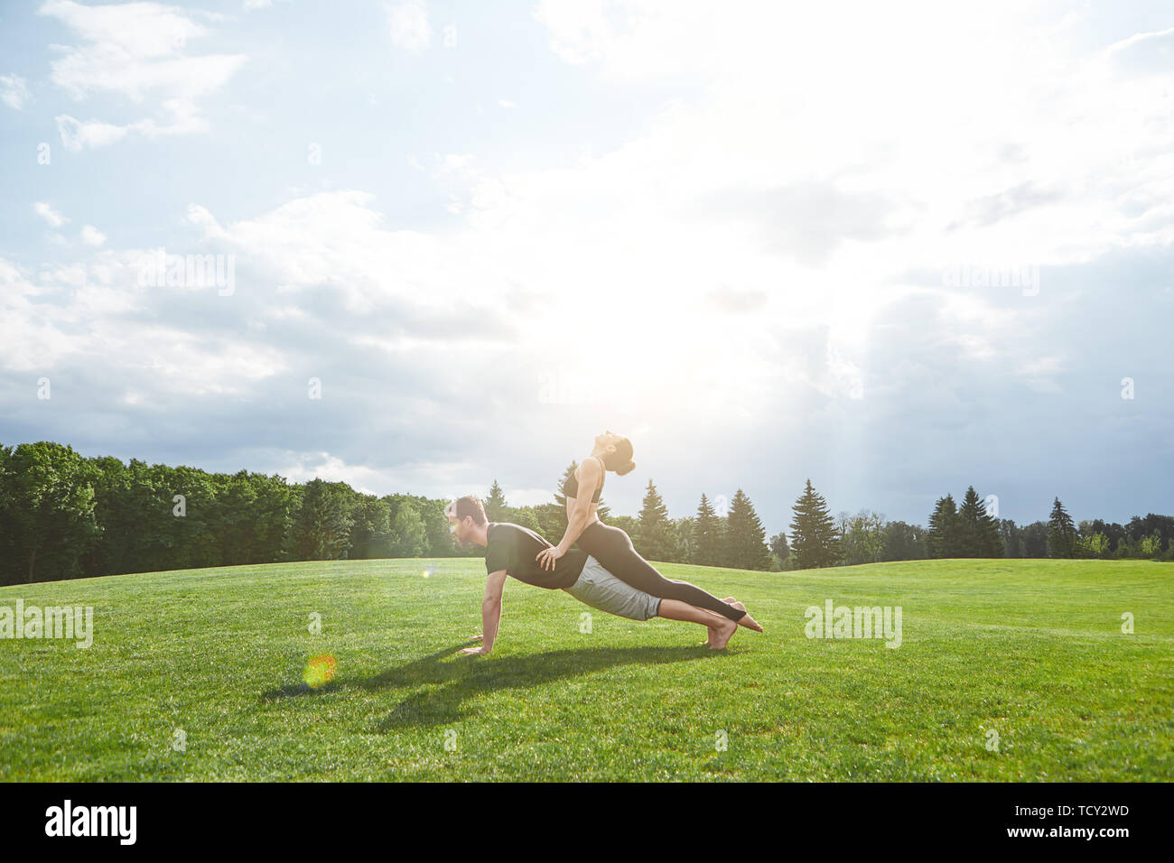 Üben acro Yoga zusammen. Starke Mann bleibt in den Liegestütz auf Gras in der Natur und Frau ist Yoga Asana durch lehnte sich auf seinen Rücken. Sonnigen morgen Stockfoto