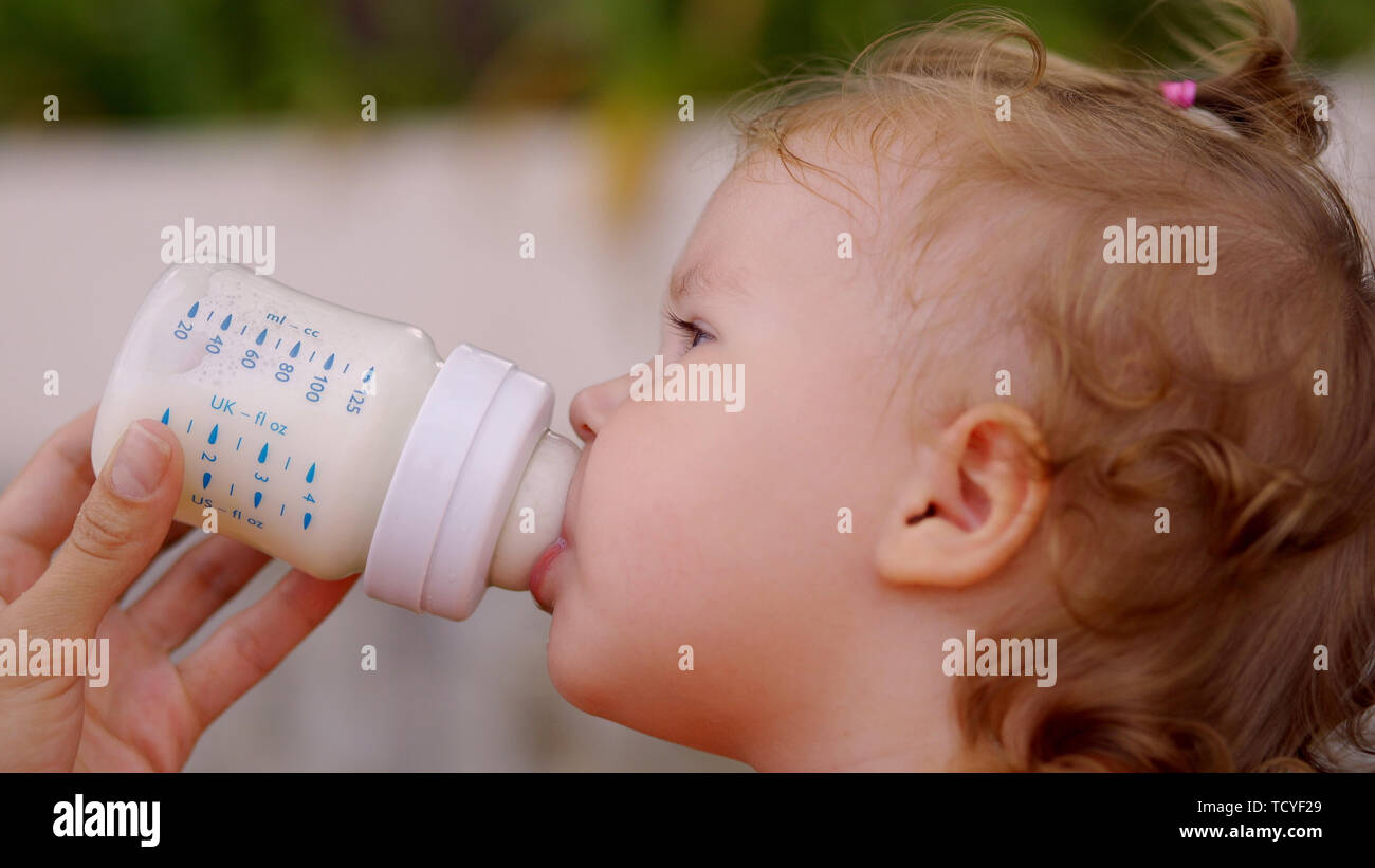 Wenig schöne Mädchen mit blauen Augen trinkt Milch aus der Flasche. Das Kind hält die Flasche selbst Stockfoto