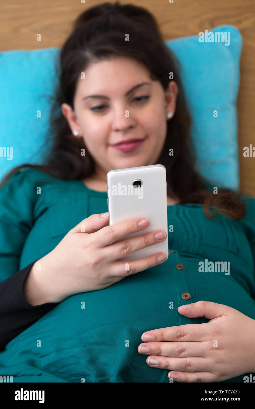Glücklich schwanger Frau sitzt auf dem Bett mit ihrem Smartphone Stockfoto
