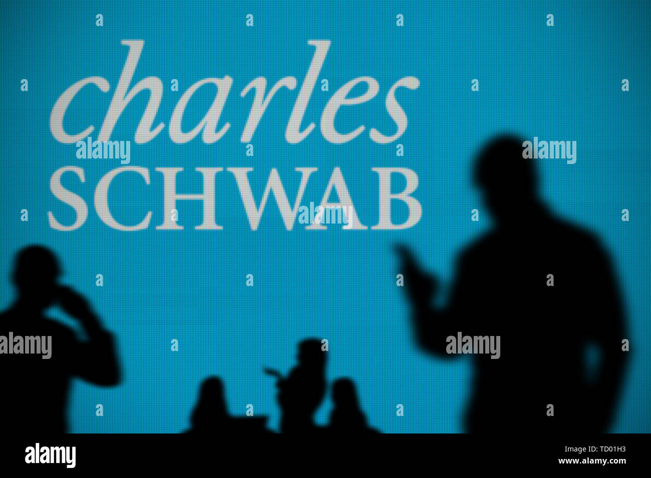 Die Charles Schwab Logo ist auf einen LED-Bildschirm im Hintergrund, während eine Silhouette Person ein Smartphone verwendet im Vordergrund (nur redaktionelle Nutzung) Stockfoto