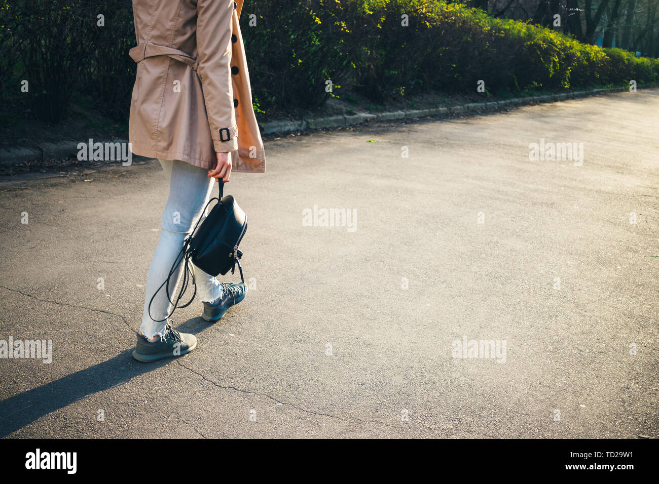 Ansicht der Rückseite des junge Frau mit Rucksack in der Hand tragen beige Mantel und graue Jeans durch den Park flanieren im Frühjahr. In der Nähe der weiblichen Beine Stockfoto