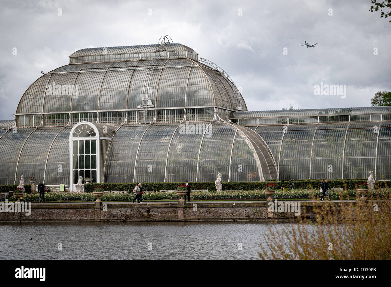 Ein Teil der Palm House mit Touristen und ein Flugzeug in einem bewölkten Himmel in Kew Gardens. Stockfoto