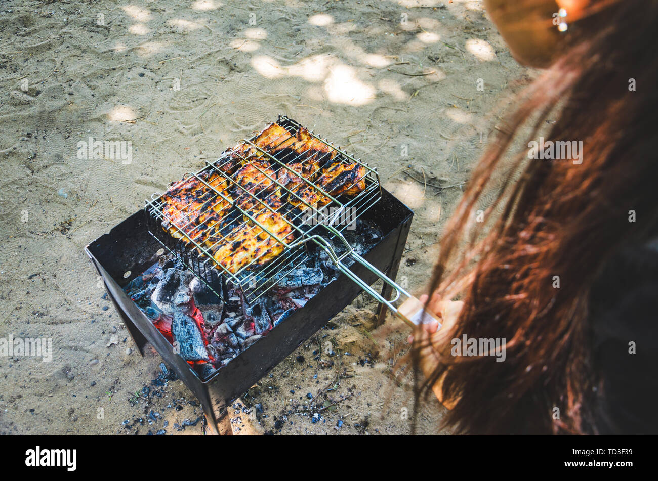 Frau mit langen dunklen Haaren halten Sie ein Raster von Grill mit Hühnerfleisch über glühende Kohle. Stockfoto