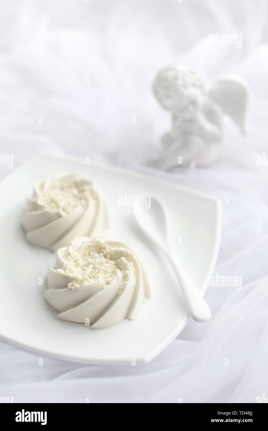Luftige dessert Panna cotta Mousse mit weißer Schokolade und Vanille auf die Oberseite, auf weißer Keramik Teller und Löffel aus Keramik und Porzellan Engel Statuette. Schwarzweiß Stockfoto