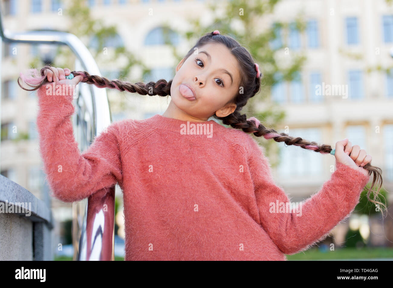 Spaß mit ihren Haaren. Funny Girl holding Haar Zöpfe im städtischen Hintergrund. Kleines süßes Mädchen mit langen brünetten Haar zeigt Zunge outdoor. Wenig Haar Modell mit verrückt aussehen. Stockfoto