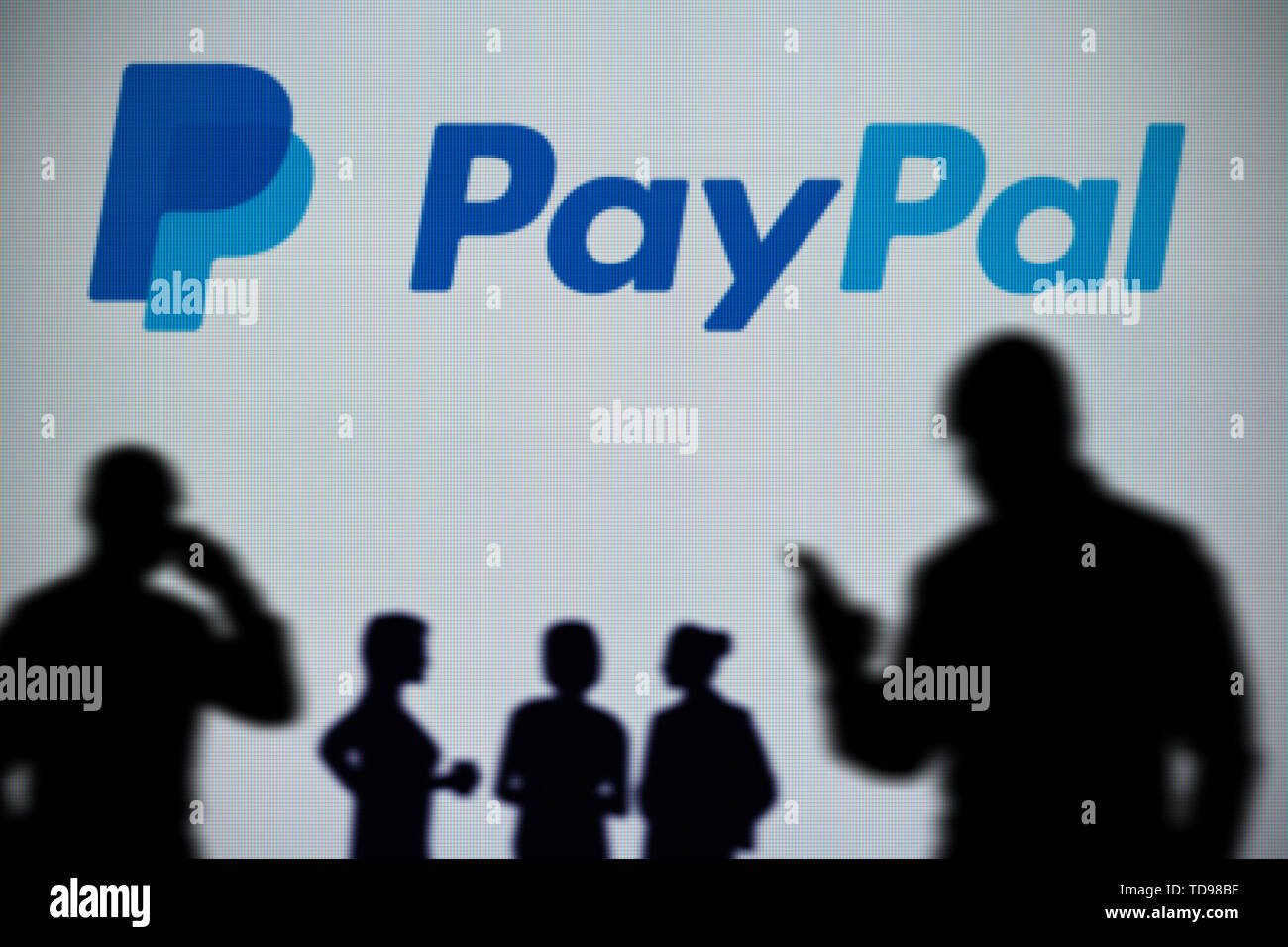 Das PayPal Logo auf einer LED-Bildschirm im Hintergrund, während eine Silhouette Person ein Smartphone verwendet im Vordergrund (nur redaktionelle Nutzung) Stockfoto