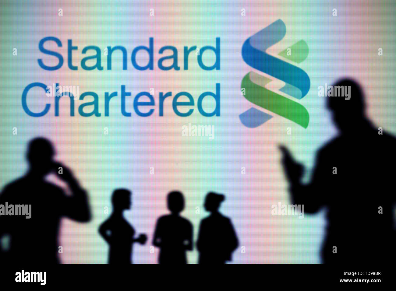 Die Standard Chartered Logo ist auf einen LED-Bildschirm im Hintergrund, während eine Silhouette Person ein Smartphone verwendet (nur redaktionelle Nutzung) Stockfoto