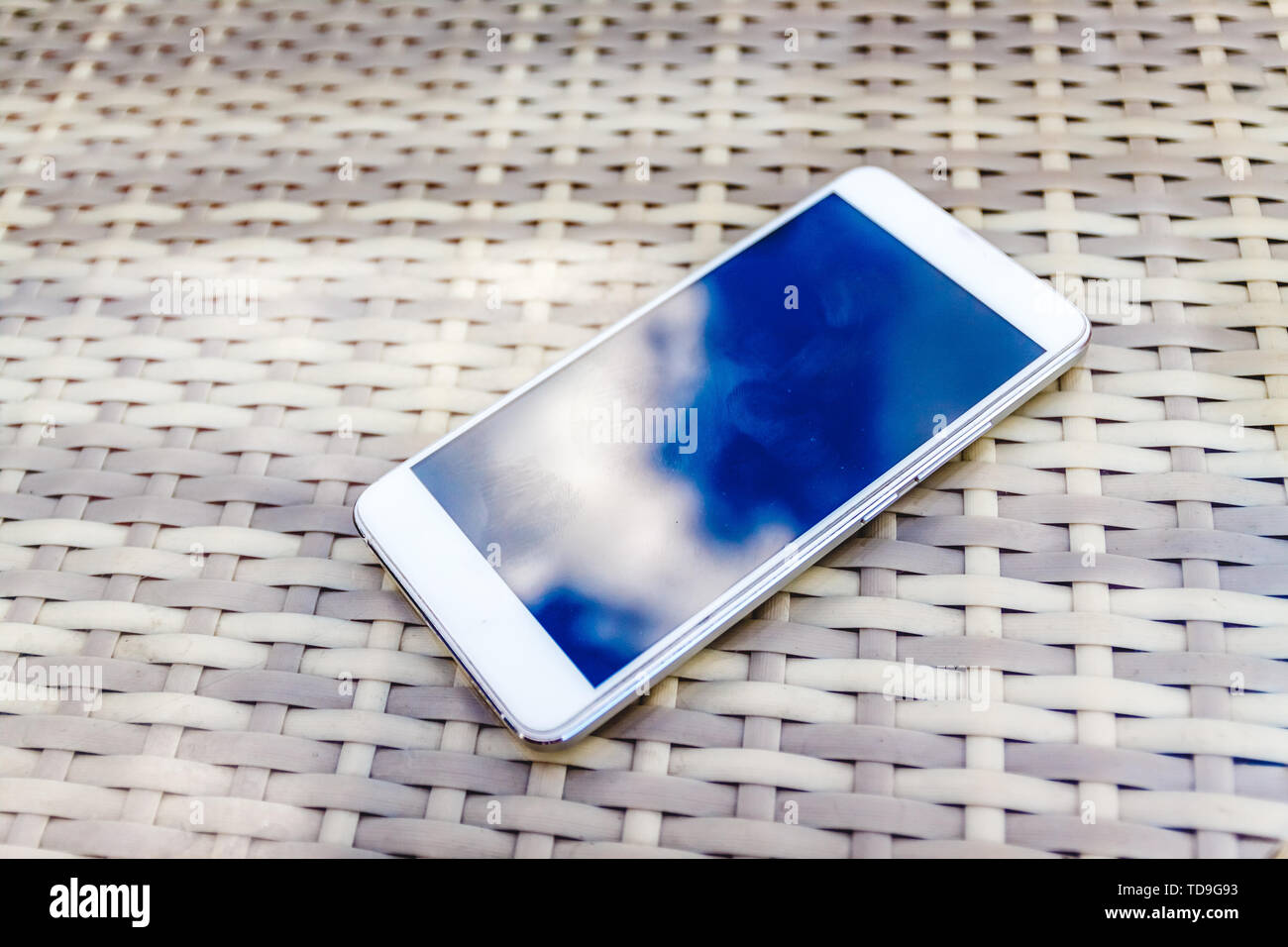 Reflexion der blauen Himmel mit Wolken im Telefon - Smartphone, das auf einem hellen Rattanmöbeln Tabelle vor dem Hintergrund liegt. Blur und Abstraktion Stockfoto