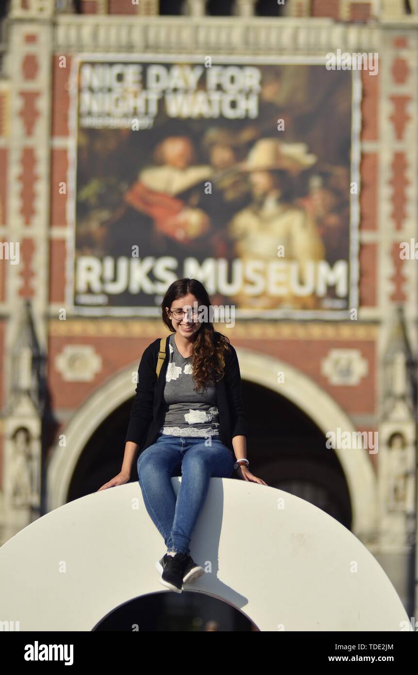 Eine junge Frau sitzt auf das Iamsterdam Zeichen vor dem Rijksmuseum, unter dem Herbst Sonnenschein im Museumsviertel Stockfoto