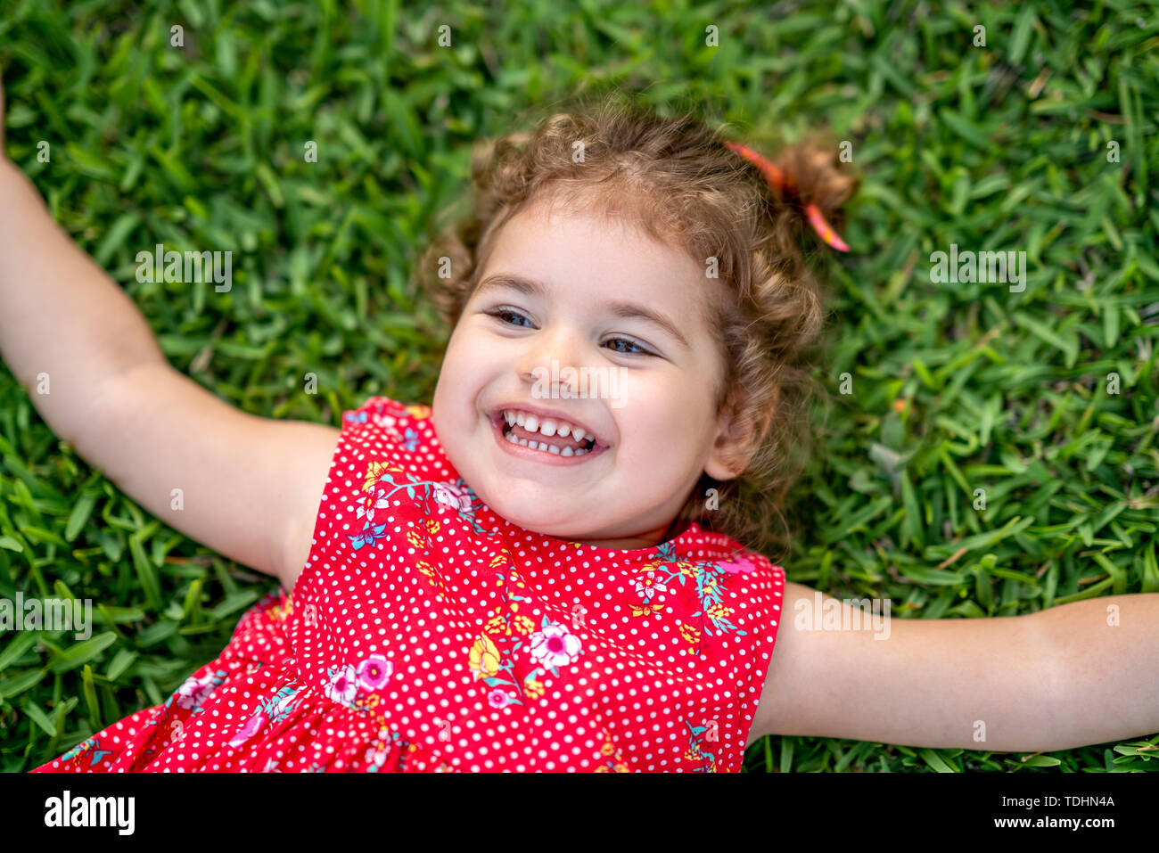 Glücklich lächelnde wenig Kleinkind Mädchen mit auf Gras im Park mit roten Kleid. Stockfoto