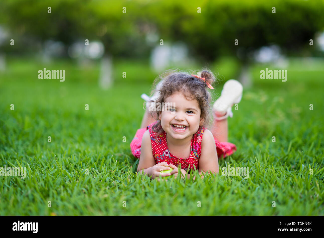 Glücklich lächelnde wenig Kleinkind Mädchen mit auf Gras im Park mit roten Kleid. Stockfoto