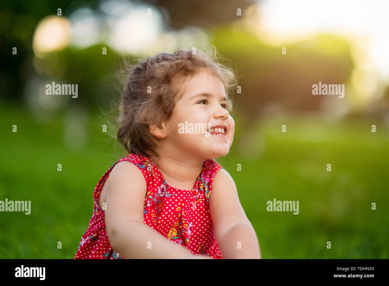 Glücklich lächelnde Mädchen sitzen auf Gras im Park mit roten Kleid. Stockfoto
