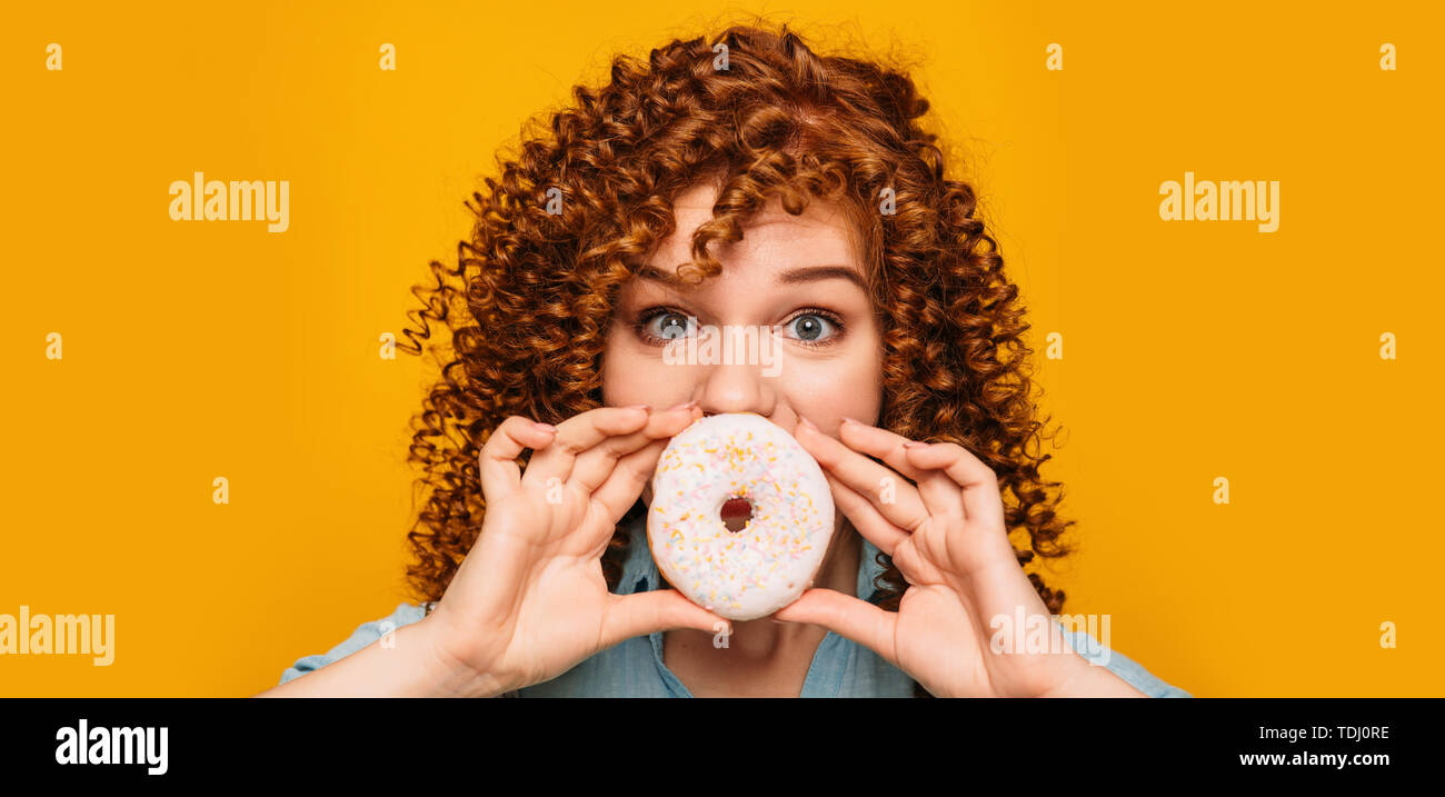 Curly rothaarige Frau mit Donut in der Nähe von Mund mit Überraschung emotionale Gesicht auf gelbem Hintergrund Stockfoto