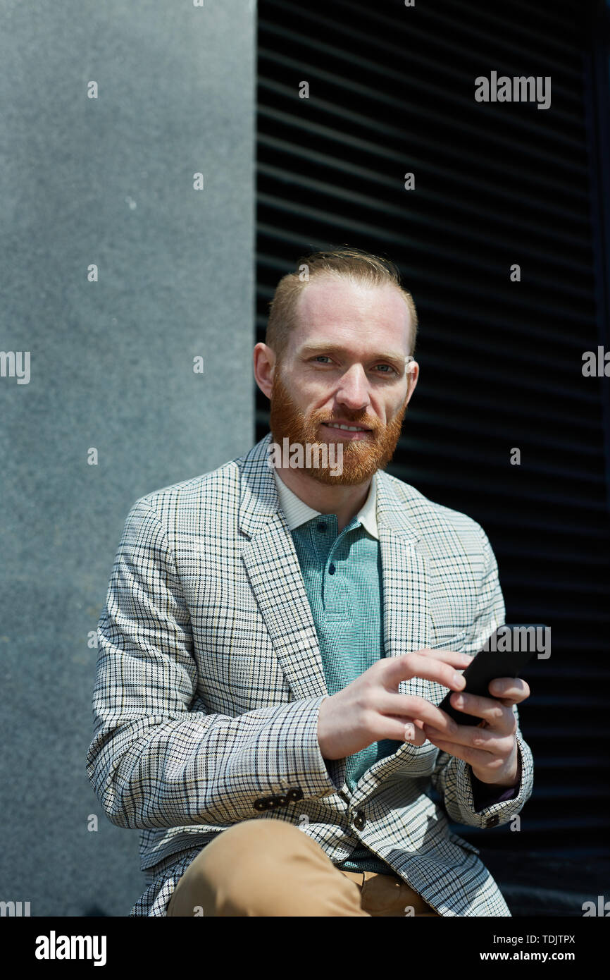 Lächelnd rotbärtigen Manager in stilvolle Jacke sitzen draußen und sms sms auf Smartphone, während bei Camera suchen Stockfoto