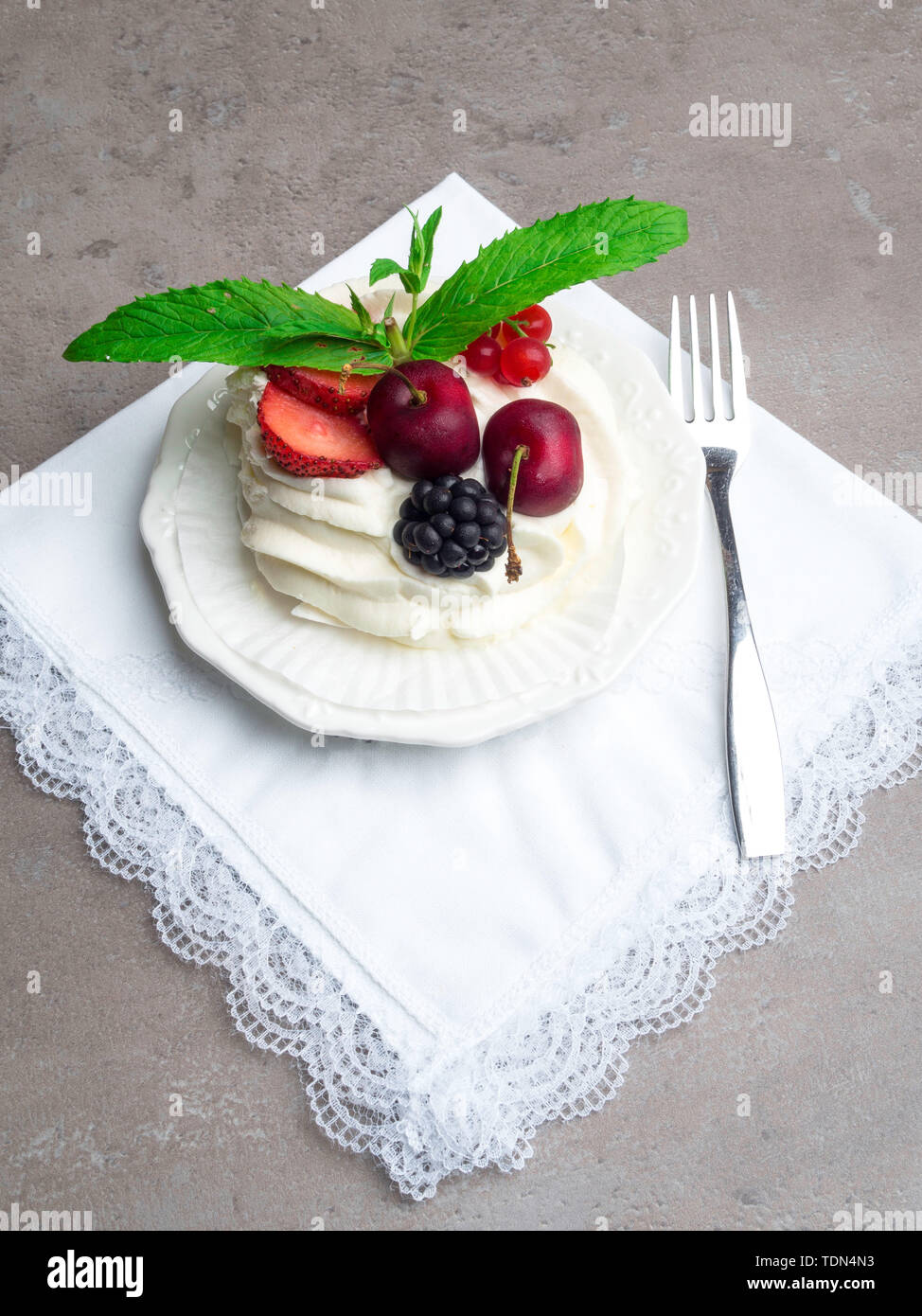 Rustikale Pavlova Kuchen mit frischen Erdbeeren, Kirschen, Blackberrys, Minze und Schlagsahne auf Beton grau hinterlegt. Stockfoto