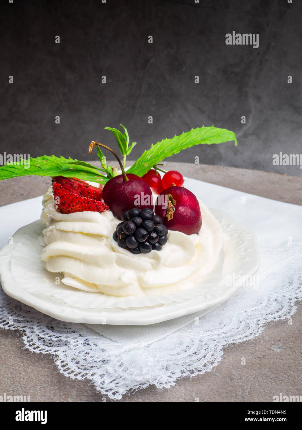 Rustikale Pavlova Kuchen mit frischen Erdbeeren, Kirschen, Blackberrys, Minze und Schlagsahne auf Beton grau hinterlegt. Stockfoto