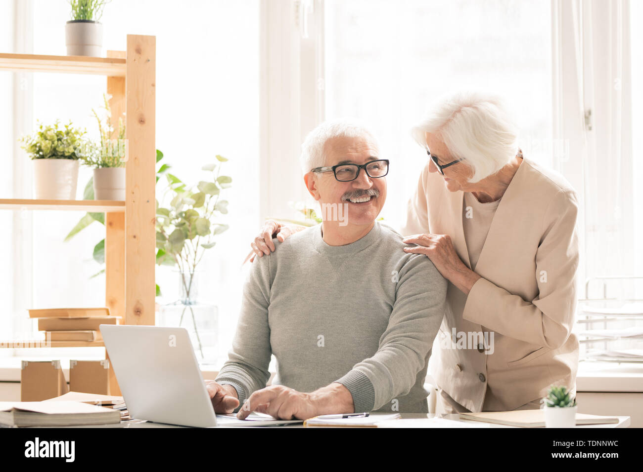 Gerne älterer Mann in der casualwear sitzen Schreibtisch vor Laptop, während im Gespräch mit seiner Frau neben ihm stehen Stockfoto