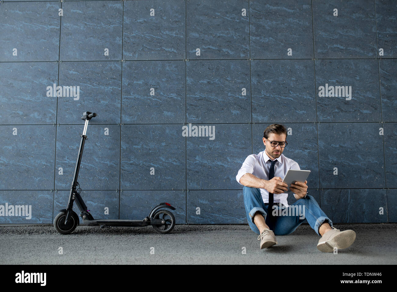 Junge Unternehmer in Smart Casual Wear entspannt sitzen durch die Wand von Gebäude und Blättern in digitalen Tablet Stockfoto