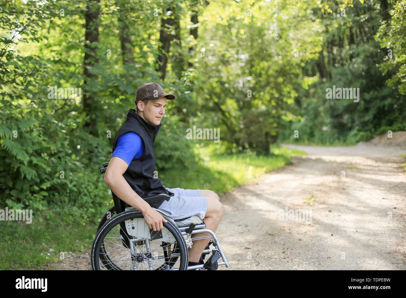 Glücklich und junge behinderte Mann sitzt auf einem Rollstuhl in der Natur drehen die Räder auf einer gehenden Straße an einem sonnigen Tag Stockfoto