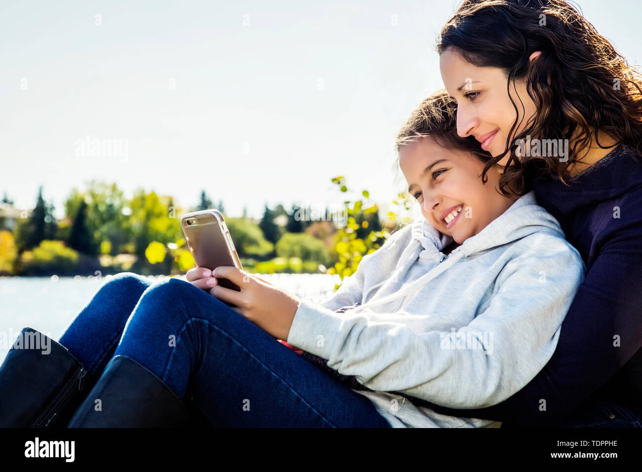 Eine Mutter und ihre Tochter, ein Selbstportrait und die Überprüfung der sozialen Medien, während an einem See in einem Stadtpark an einem warmen Herbst Tag ruhen Stockfoto