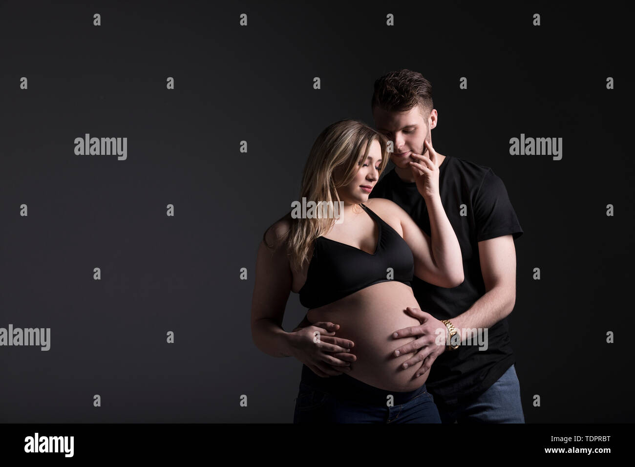 Eine junge werdende Paar unten betrachten Ihr Baby während der Vater ihren Bauch in einem Studio auf einem dunklen Hintergrund hält: Edmonton, Alberta, Kanada Stockfoto