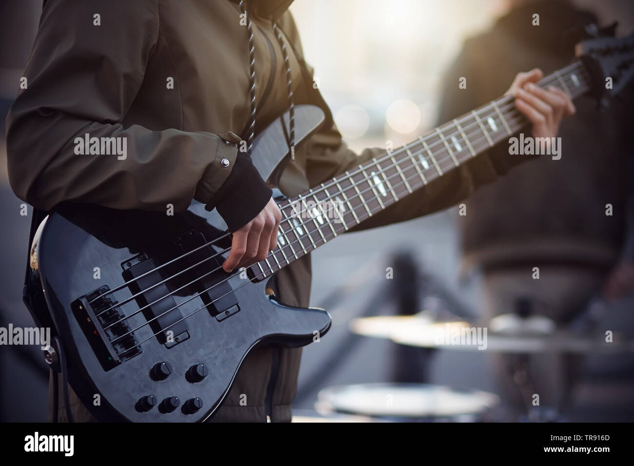 Ein Straßenmusiker, in einer dunklen Jacke mit gestreiften Schnürsenkel gekleidet, spielt ein Rhythmus, der auf einem 5-String E-Gitarre, stand am Abend auf dem Platz Stockfoto