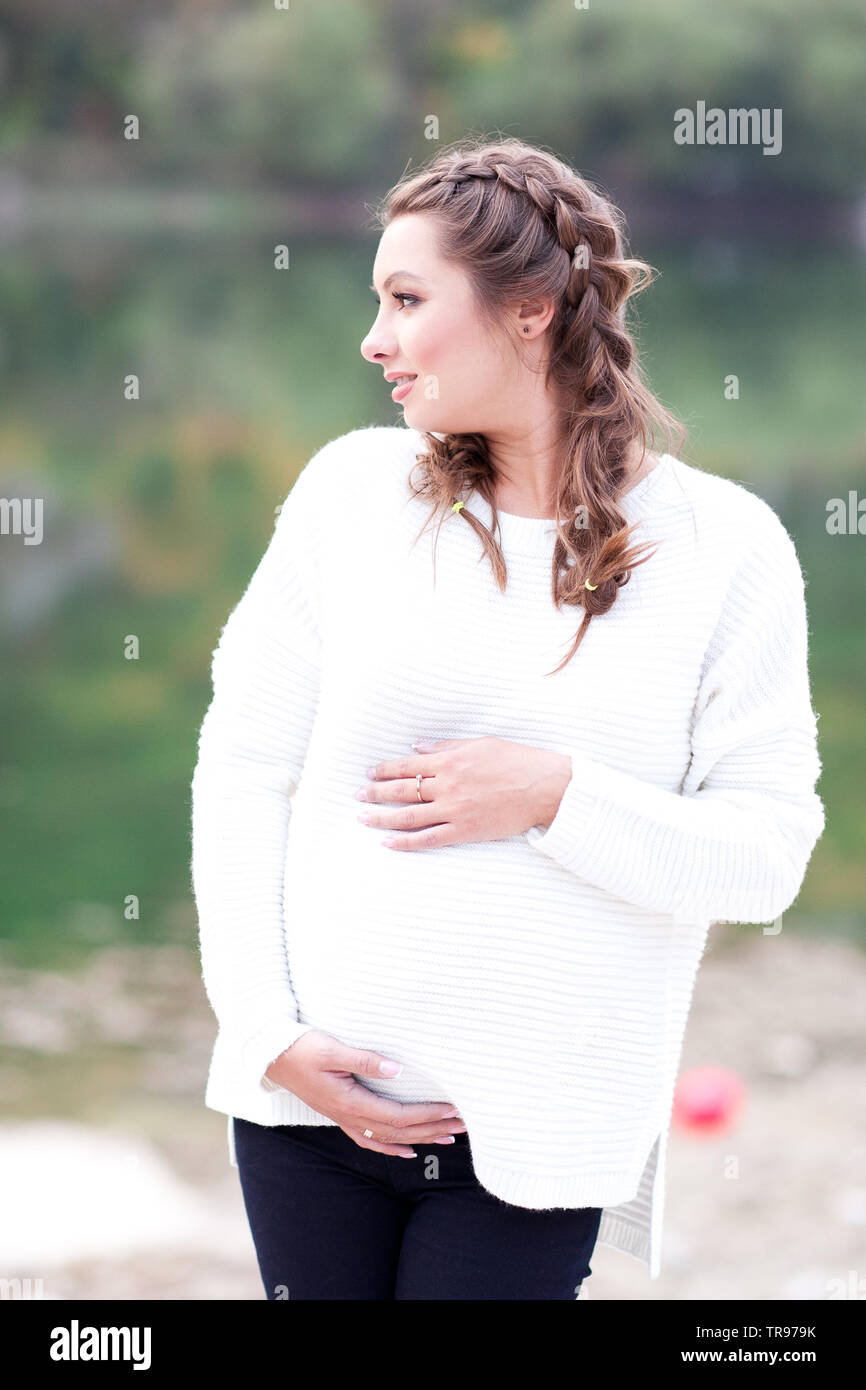 Lächelnd schwangere Frau mit Bauch. Tragen weiße gestrickte Pullover  posiert im Freien. Weg schauen. Die Mutterschaft Stockfotografie - Alamy