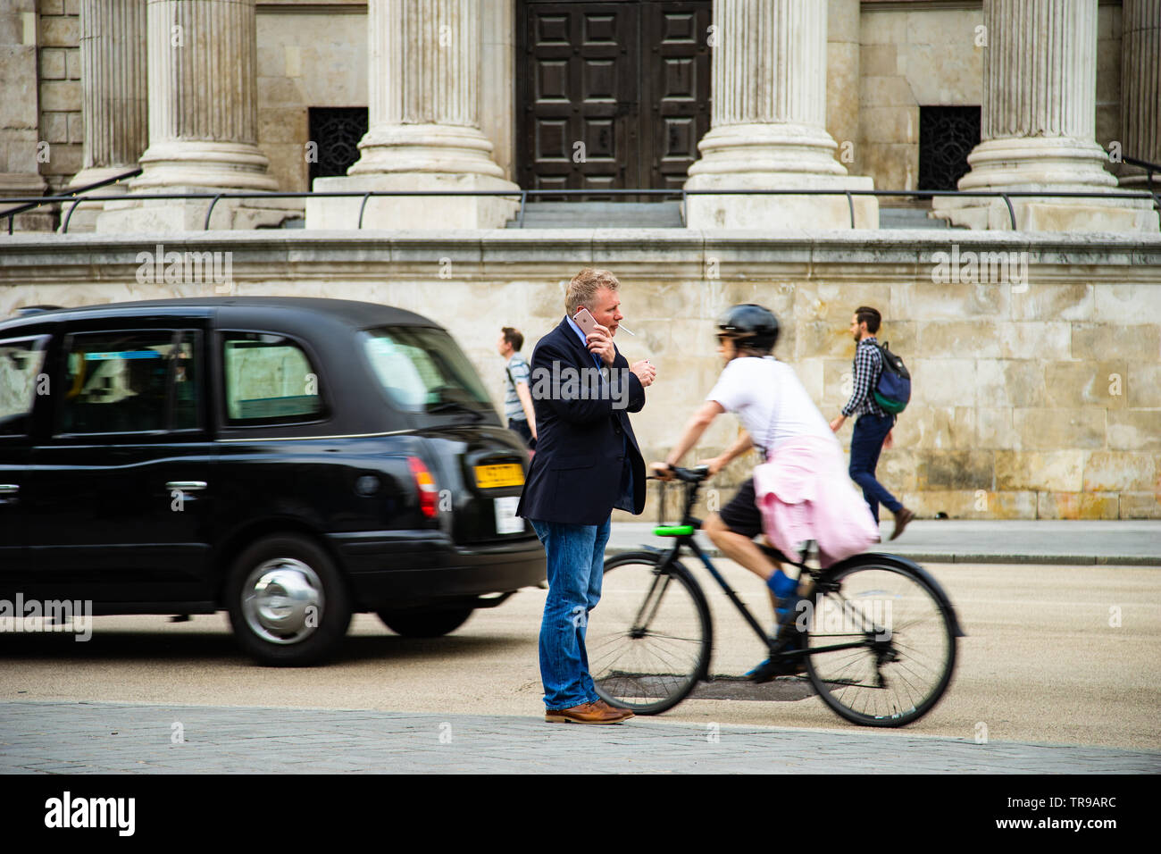 Ein Mann steht inmitten des Chaos in London, so leuchten seine Zigarette während des Sprechens auf seinem iPhone als Radfahrer und black cab vorbei Stockfoto