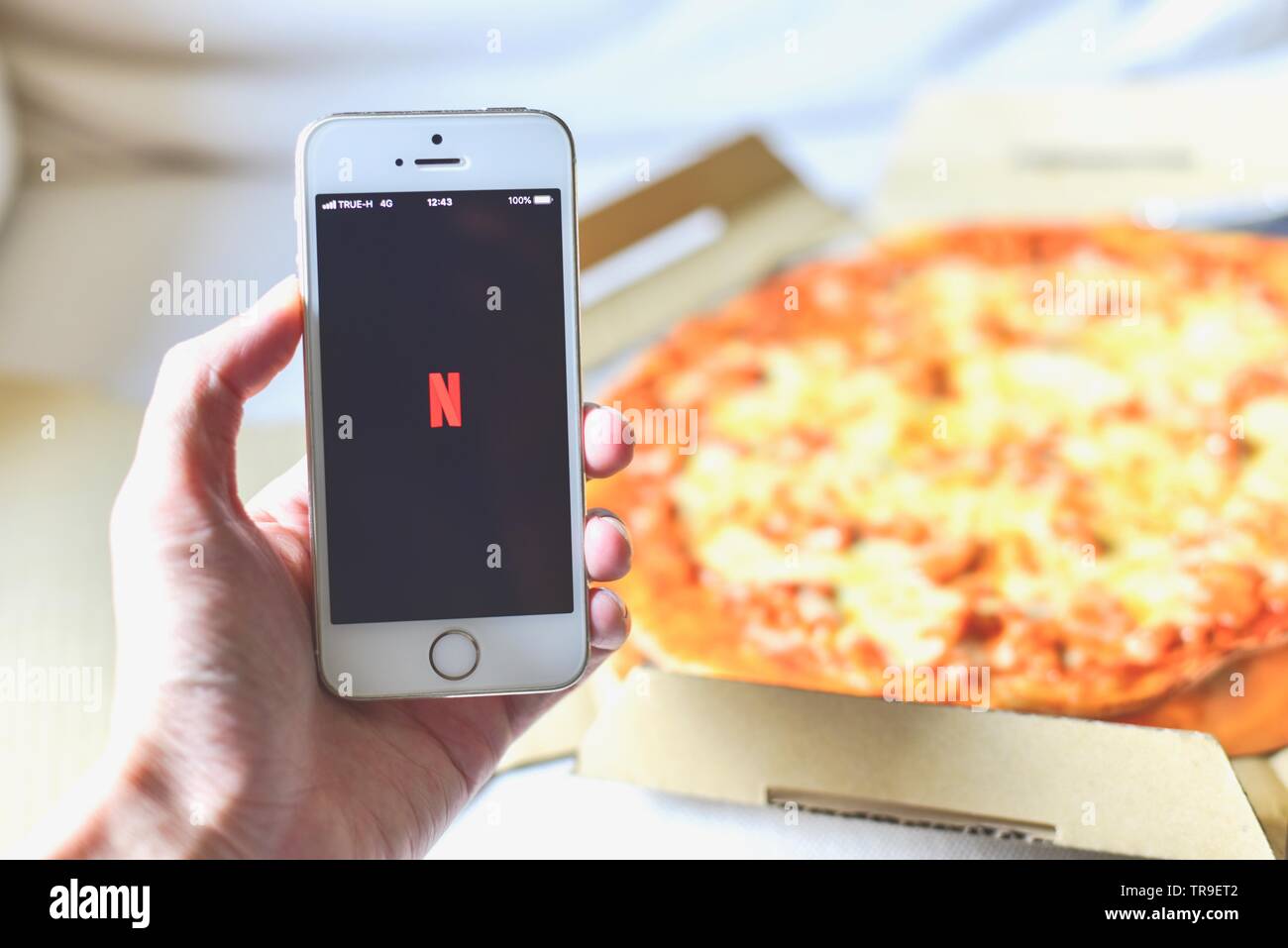 Hand mit einem Smartphone mit Netflix Anwendung auf dem Bildschirm und Pizza im Hintergrund Stockfoto